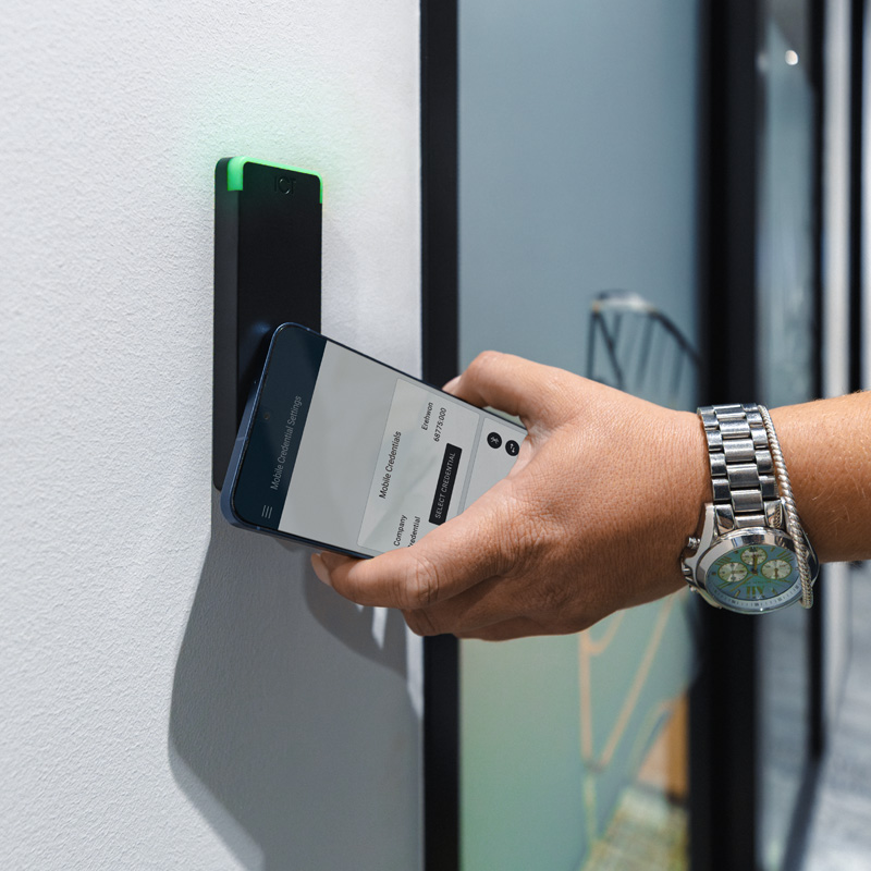 A person tapping a smartphone against an ICT reader to unlock a glass door