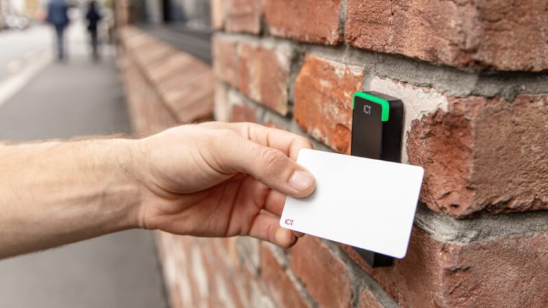 A hand tapping a physical ICT access card against a wall-mounted reader